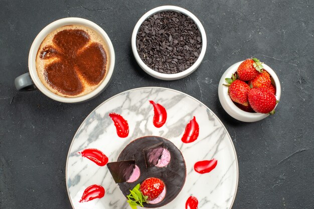 Cheesecake alla fragola vista dall'alto su ciotole di piatto ovale bianco con fragole e cioccolato una tazza di caffè su sfondo scuro