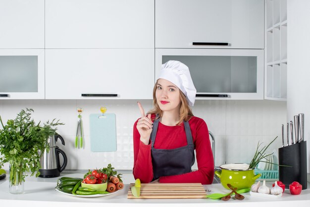 Chef donna vista frontale in uniforme in piedi dietro il tavolo della cucina che punta al dito