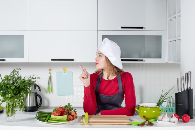 Chef donna vista frontale in uniforme in piedi dietro il tavolo della cucina guardando a sinistra