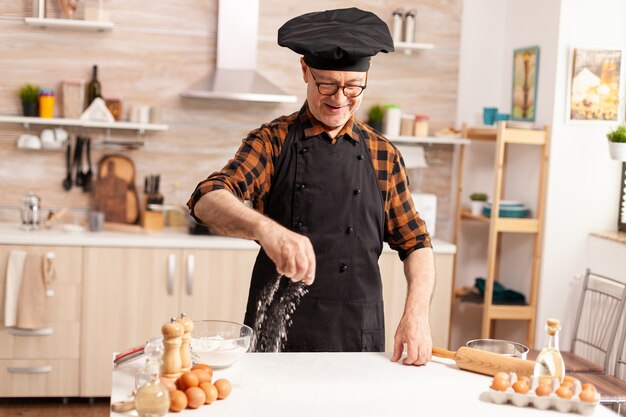 Chef in pensione nella cucina di casa che indossa un grembiule mentre cosparge l'ingrediente sul tavolo per un gustoso