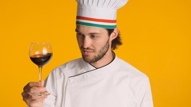 Chef italiano vestito in uniforme tenendo un bicchiere di vino rosso su sfondo colorato Sommelier uomo degustazione di buon vino