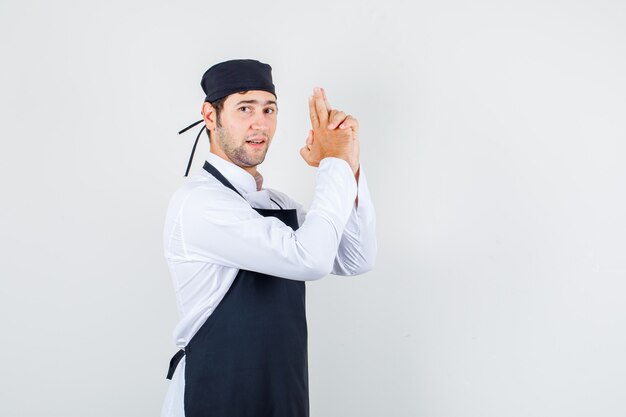 Chef maschio che fa il gesto della pistola di tiro in uniforme, grembiule e guardando fiducioso, vista frontale.