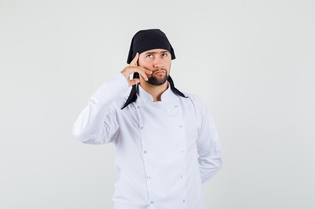 Chef maschio in uniforme bianca alzando lo sguardo con il dito sulle tempie e guardando premuroso, vista frontale.