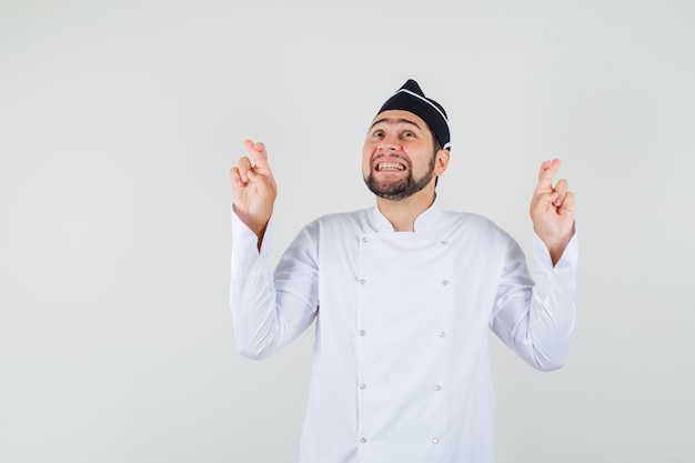 Chef maschio in uniforme bianca alzando lo sguardo con le dita incrociate e guardando allegro, vista frontale.