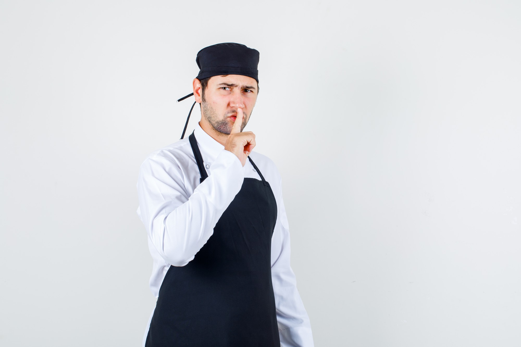 Chef maschio in uniforme, grembiule che mostra gesto di silenzio mentre accigliato, vista frontale.