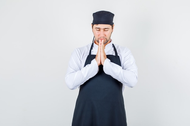 Chef maschio in uniforme, grembiule mano nella mano nel gesto di preghiera e guardando in silenzio, vista frontale.