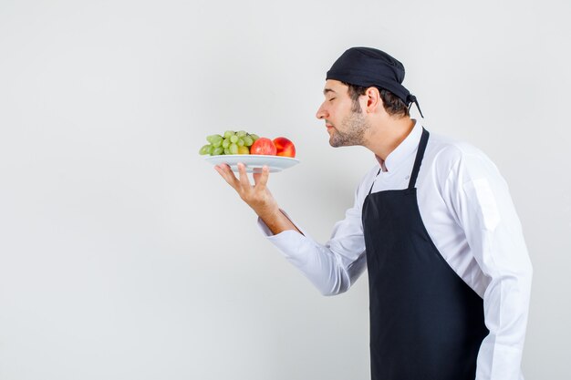 Chef maschio in uniforme, grembiule sentente l'odore di frutta nel piatto, vista frontale.