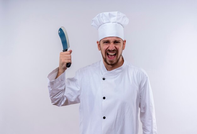 Chef maschio professionista cuoco in uniforme bianca e cappello da cuoco che oscilla un coltello affilato con espressione aggressiva in piedi su sfondo bianco