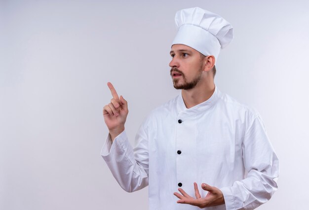 Chef maschio professionista cuoco in uniforme bianca e cappello da cuoco che punta il dito verso l'alto guardando da parte pensando in piedi su sfondo bianco