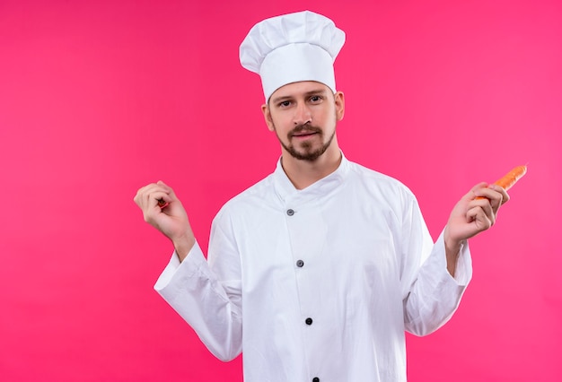 Chef maschio professionista cuoco in uniforme bianca e cappello da cuoco che tiene la carota che guarda l'obbiettivo con il sorriso sul viso in piedi su sfondo rosa