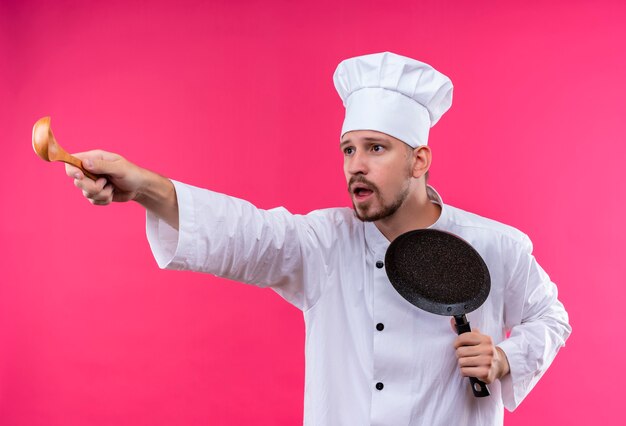 Chef maschio professionista cuoco in uniforme bianca e cappello da cuoco che tiene una padella che punta con un cucchiaio di legno di lato guardando sorpreso e preoccupato in piedi su sfondo rosa