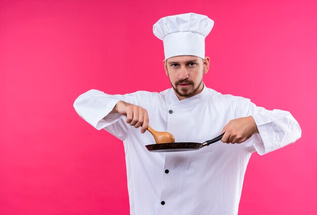 Chef maschio professionista cuoco in uniforme bianca e cappello da cuoco che tiene una padella e un cucchiaio di legno guardando fiducioso in piedi su sfondo rosa