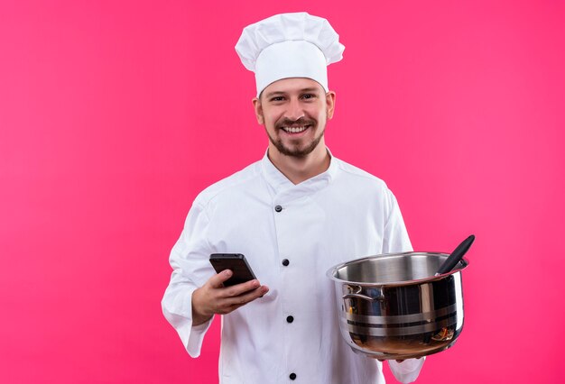 Chef maschio professionista cuoco in uniforme bianca e cappello da cuoco che tiene una padella vuota e un telefono cellulare che sorride allegramente in piedi su sfondo rosa