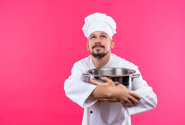 Chef maschio professionista cuoco in uniforme bianca e cappello da cuoco che tiene una pentola vuota che osserva in su con sguardo sognante in piedi su sfondo rosa