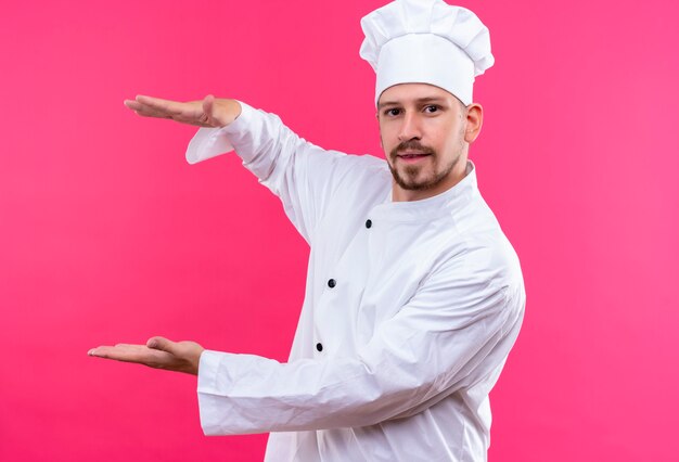 Chef maschio professionista cuoco in uniforme bianca e cappello da cuoco gesticolando con le mani che mostrano le dimensioni, simbolo di misura su sfondo rosa