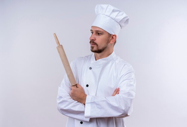 Chef maschio professionista cuoco in uniforme bianca e cappello da cuoco in piedi con le braccia incrociate tenendo il mattarello che guarda da parte con espressione fiduciosa su sfondo bianco