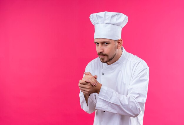 Chef maschio professionista cuoco in uniforme bianca e cappello da cuoco strofinando le mani guardando la telecamera furbescamente in piedi su sfondo rosa