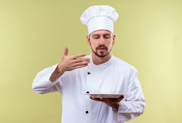 Chef maschio professionista cuoco in uniforme bianca e cappello da cuoco tenendo la piastra inala l'odore gradevole del cibo in piedi su sfondo verde