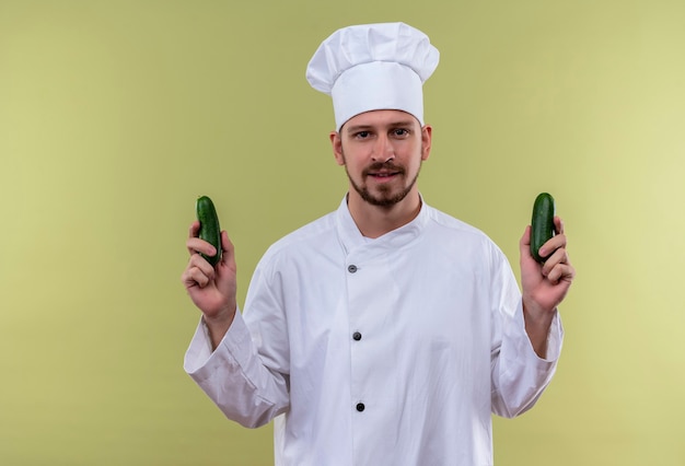 Chef maschio professionista soddisfatto cucinare in uniforme bianca e cappello da cuoco che tiene i cetrioli freschi in piedi su sfondo verde