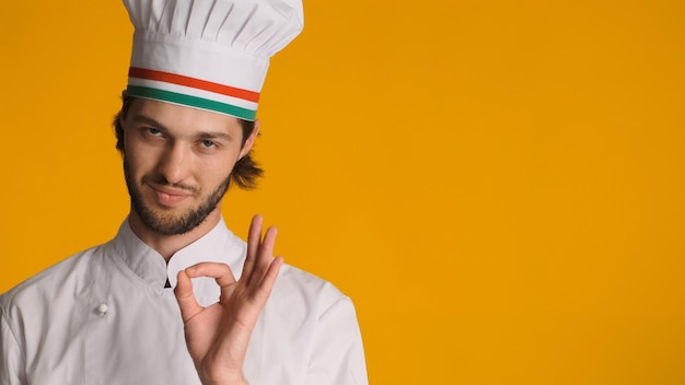 Chef maschio sicuro in uniforme in piedi vicino allo spazio per la pubblicità che mostra segno ok su sfondo arancione Uomo in cappello da chef che mostra gesto approvato