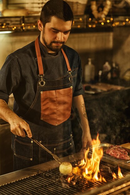 Chef professionista che cucina cipolla verdure e carne alla griglia con fuoco e fumo bell'uomo concentrato sulla preparazione del cibo sullo sfondo della moderna cucina del ristorante