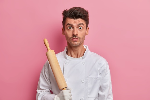 Chef sorpreso con utensile da cucina, vestito in uniforme bianca, panettiere impegnato tiene il mattarello