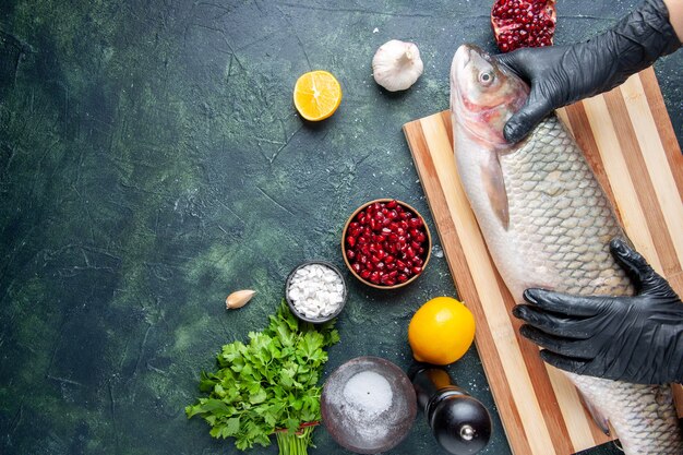 Chef vista dall'alto con guanti neri che tengono pesce crudo su tavola di legno macinapepe semi di melograno in una ciotola sullo spazio libero del tavolo