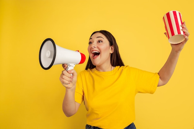 Chiamando con la bocca aperta e popcorn. Donna caucasica sulla parete gialla. Bellissima modella bruna femminile in stile casual. Concetto di emozioni umane, espressione facciale, vendite, annuncio, copyspace.