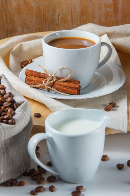 Chicchi di caffè in un sacco, latte, cannella secca e una tazza di caffè su una piattaforma e un tavolo bianco. vista laterale.