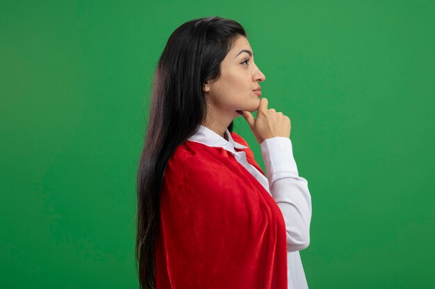 Chiedendosi giovane supereroe caucasico ragazza in piedi in vista di profilo mettendo la mano sulla mascella guardando dritto isolato su sfondo verde con spazio di copia