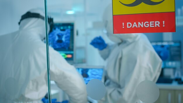 Chimici in tuta che cercano di sviluppare un vaccino usando un tablet in piedi dietro la parete di vetro che lavora in un'area pericolosa del laboratorio