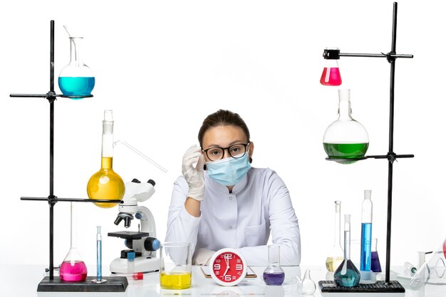 Chimico femminile di vista frontale in vestito medico con la maschera che si siede con le soluzioni sul laboratorio covid di chimica del virus della spruzzata del fondo bianco