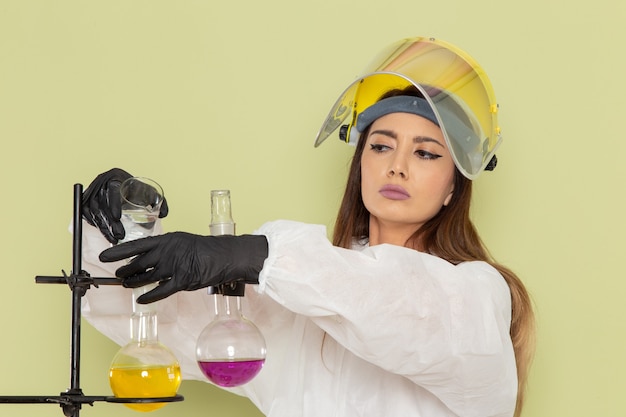 Chimico femminile giovane vista frontale in tuta protettiva speciale che lavora con diverse soluzioni sulla scienza femminile di chimica del laboratorio chimico di lavoro della parete verde