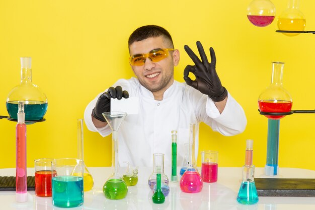 Chimico maschio giovane vista frontale in abito bianco davanti al tavolo con soluzioni colorate che tiene carta bianca sorridente sulla chimica del laboratorio di lavoro di scienza parete gialla