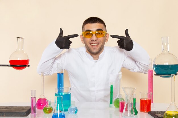 Chimico maschio giovane vista frontale in abito speciale bianco davanti al tavolo con soluzioni colorate sorridente sul muro crema laboratorio chimica scienza lavoro prodotti chimici