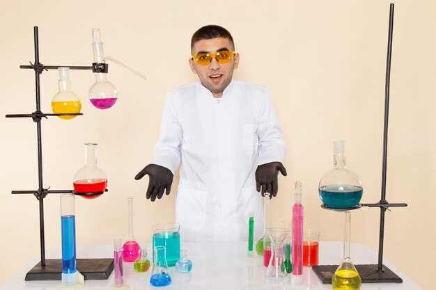 Chimico maschio giovane vista frontale in abito speciale bianco davanti al tavolo con soluzioni in posa sulla scienza chimica di laboratorio parete crema