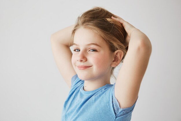 Chiuda in su della bambina allegra in maglietta blu che osserva, sorridendo brillantemente e che tiene i capelli lunghi biondi con le mani. Copia spazio.