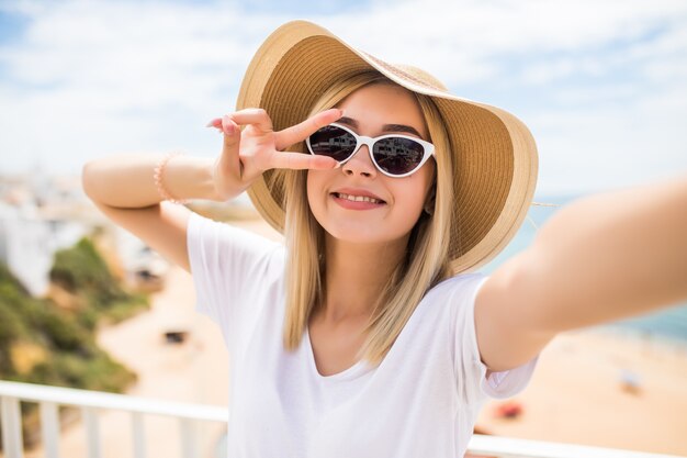 Chiuda in su della bella ragazza giovane in cappello estivo prendendo un selfie e mostrando il gesto di pace in spiaggia