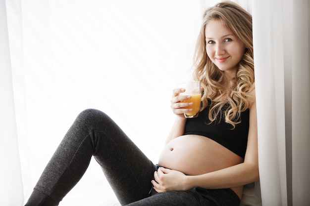 Chiuda in su della donna incinta europea felice allegra con capelli ondulati biondi in vestiti accoglienti che sorride delicatamente a porte chiuse, bevendo il succo di arancia