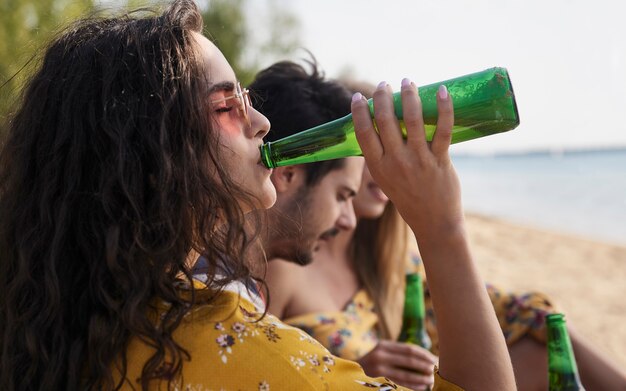 Chiuda in su della ragazza che beve birra con gli amici sulla spiaggia.