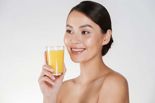 Chiuda in su della signora seminuda con pelle fresca sana e ampio sorriso che beve il succo di arancia da vetro trasparente, isolato sopra la parete bianca