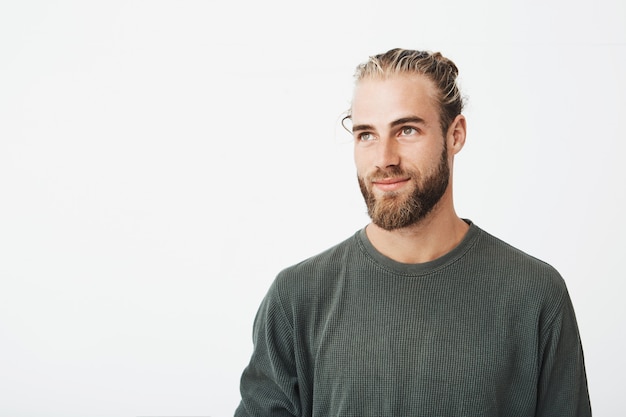 Chiuda in su di bello ragazzo virile con capelli chiari, capelli alla moda e barba in camicia grigia che sorride e che osserva da parte con l'espressione piacevole.