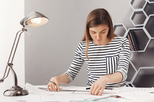 Chiuda in su di giovane progettista femminile femminile serio bello con capelli scuri lunghi in vestiti a strisce alla moda. lavorando su un nuovo progetto di squadra usando righello e penna.