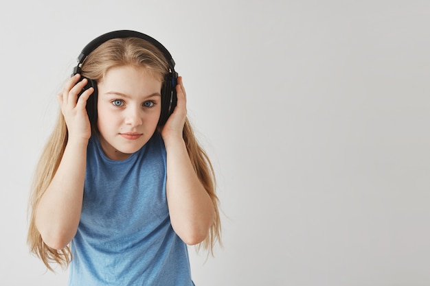 Chiuda su della bambina felice con capelli biondi in cuffie blu della tenuta della maglietta con le mani, ascoltando attentamente il testo di audizione a scuola.