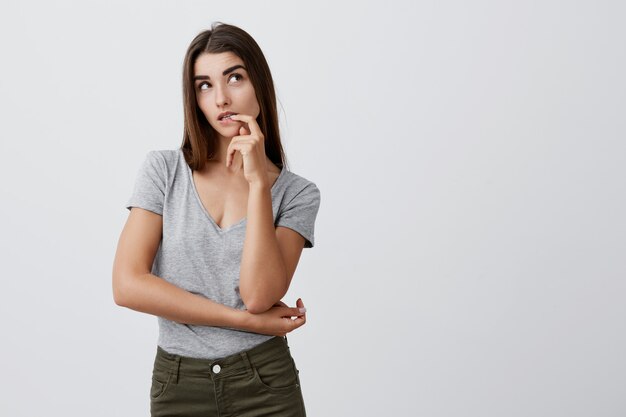 Chiuda su della ragazza caucasica seria sexy attraente con capelli lunghi scuri in camicia e jeans grigi casuali che tengono la mano vicino alla bocca, guardando da parte con l'espressione premurosa e subdola.