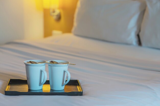 Chiuda su della tazza di caffè di benvenuto gemellata sul letto bianco nella camera di albergo - concetto di viaggio di vacanza di ospitalità dell&#39;hotel bene