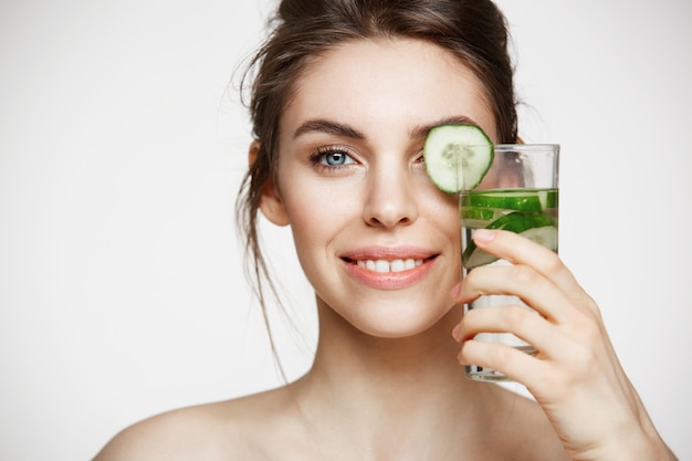 Chiuda su di bella ragazza nuda che sorride esaminando la macchina fotografica che tiene il bicchiere d'acqua con le fette del cetriolo sopra fondo bianco. Nutrizione Sana. Bellezza e cura della pelle.