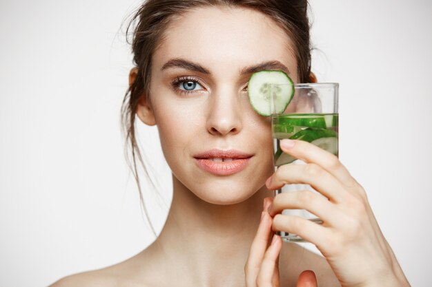 Chiuda su di bella ragazza nuda che sorride esaminando la macchina fotografica che tiene il bicchiere d'acqua con le fette del cetriolo sopra fondo bianco. Nutrizione Sana. Bellezza e cura della pelle.