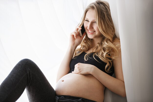 Chiuda su di giovane bella madre futura allegra con capelli leggeri lunghi in vestiti neri che si siedono sul davanzale della finestra in camera da letto