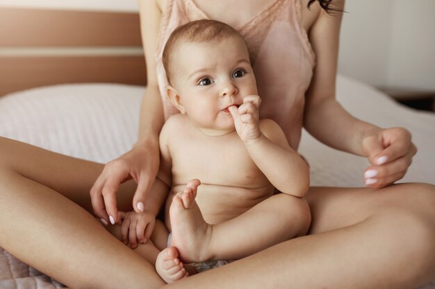 Chiuda su di giovane madre in indumenti da notte e nel suo neonato che si siedono sul letto nella mattina che sorride giocando insieme